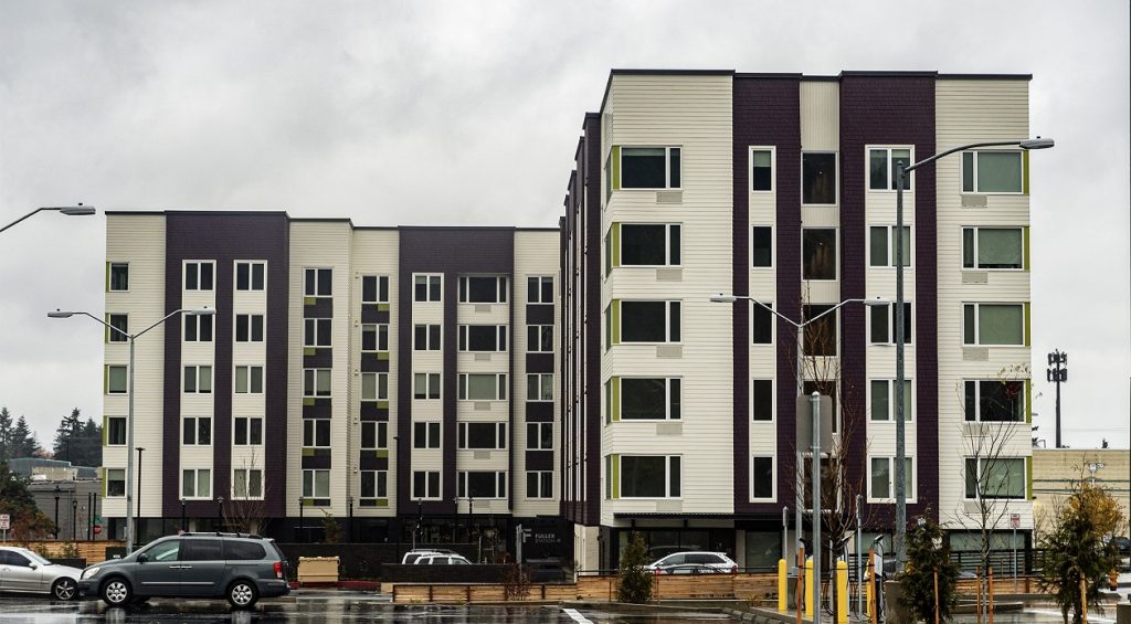 A six-story apartment complex, where one corner is closer than the other, and grey skies in the background.