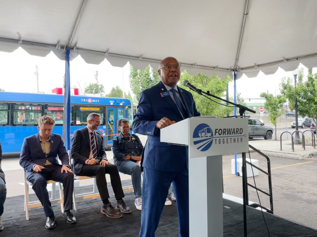 TriMet General Manager Sam Desue Jr. speaks on a stage outside the Oregon City Transit Center behind a podium labeled "Forward Together."