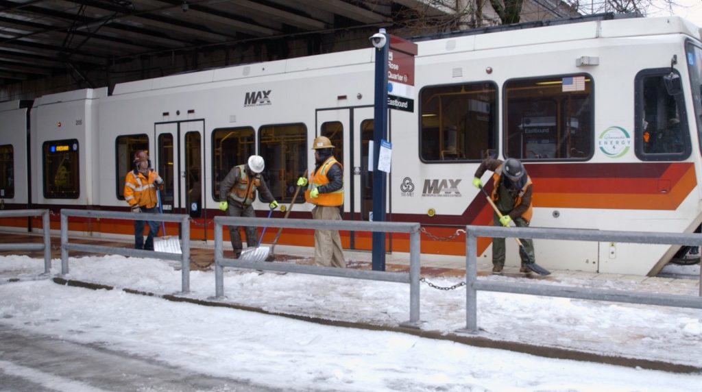 Image of TriMet MAX train at the Rose Quarter Transit Center, alongside workers shoveling snow on the platform.