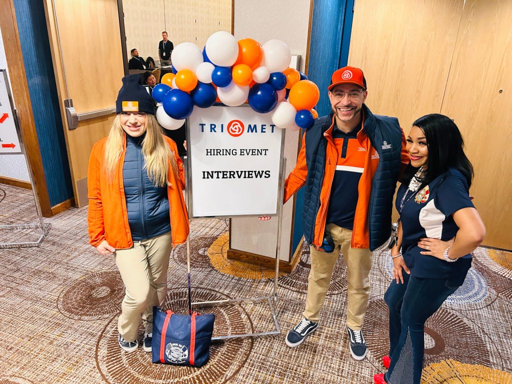 Image of three TriMet employees posing next to a sign reading "TriMet Hiring Event Interviews."