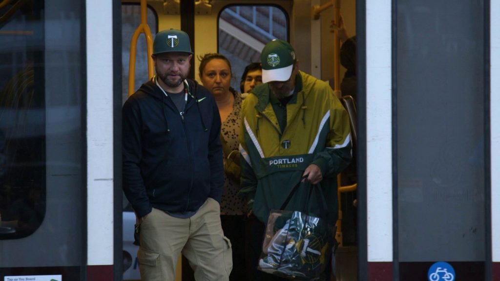 Image of four people, two of them wearing Portland Timbers caps, standing in the doorway of a TriMet MAX train.