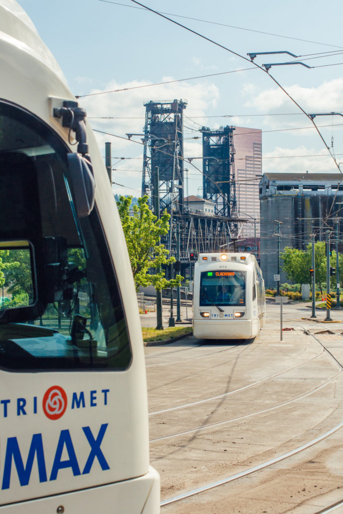 Image of two TriMet MAX trains preparing to pass near the Rose Quarter Transit Center in Northeast Portland. The Steel Bridge is visible in the background.