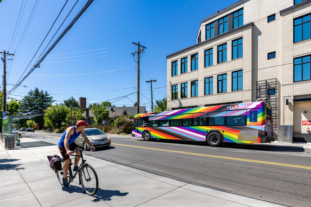 Image of TriMet's "From Progress to Beyond" artistic bus wrap by Daniel Quasar on a Portland city street. A person on a bicycle riding past the bus appears to be looking at it.
