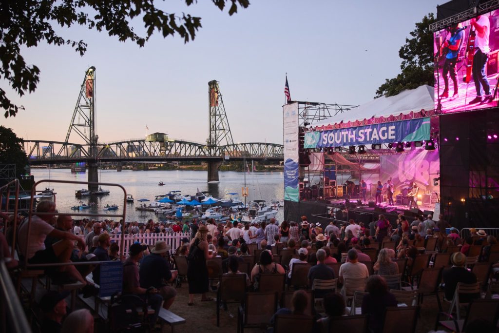 Image of a crowd listening to a concert at the Waterfront Blues Festival South Stage. The Hawthorne Bridge and boats on the Willamette River are in the background.