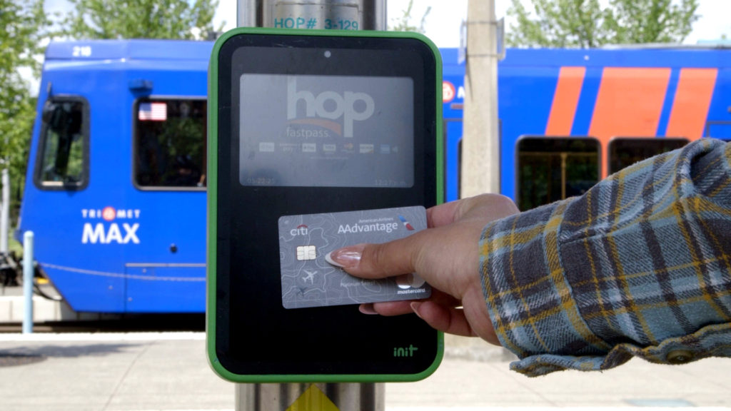 Image of a rider's hand tapping a bank card to a Hop reader at a MAX station. A MAX train is in the background.