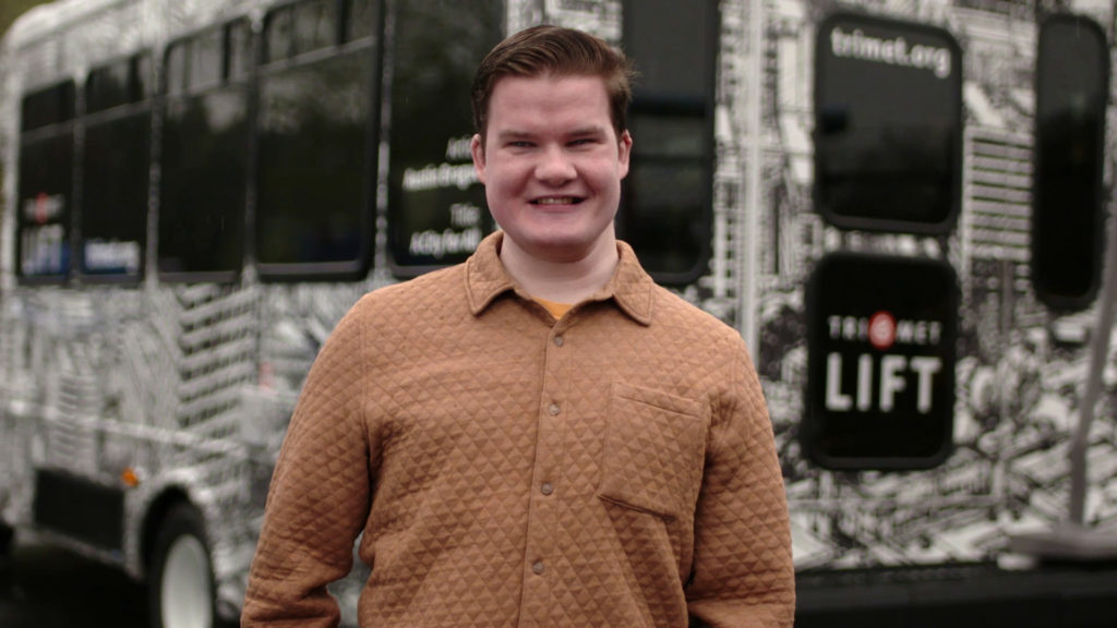 Image of Austin Brague, a young man with brown hair wearing a quilted orange shirt, in front of a TriMet LIFT bus.