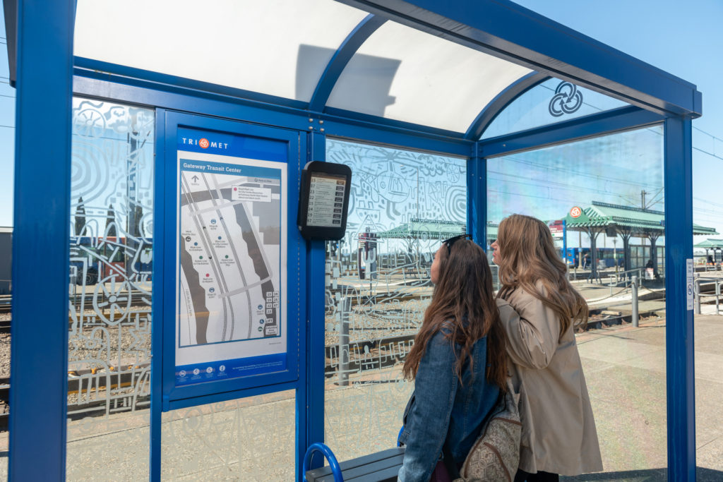 Image of two people looking at an electronic paper display with arrival times in a TriMet bus shelter at the Gateway/NE 99th Ave Transit Center.