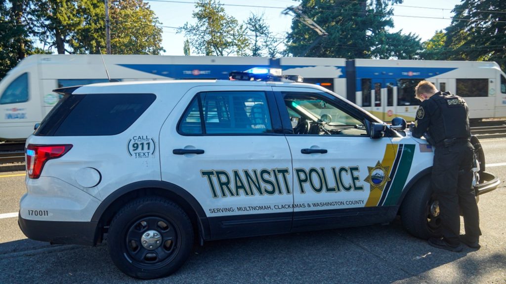 Transit Police officer next to Transit Police vehicle with a TriMet MAX train in the background. The Transit Police vehicle has text that says "Serving Multnomah, Clackamas and Washington County